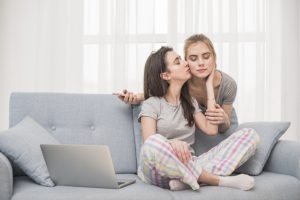lesbian-woman-sitting-on-sofa-kissing-her-girlfriend-holding-mobile-phone-in-hand_23-2148126992