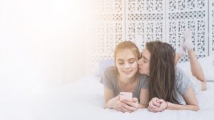 young-lesbian-woman-lying-on-bed-kissing-her-girlfriend-using-mobile-phone_23-2148126977